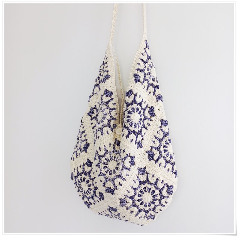 Puede incluir: Una bolsa de mano de crochet blanca con un patr&oacute;n floral azul. La bolsa tiene una correa larga y est&aacute; hecha de un hilo suave y texturizado.
