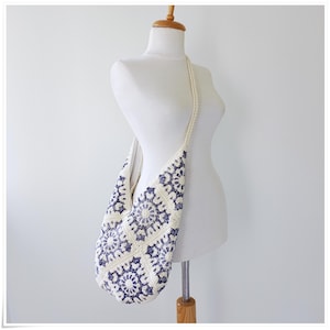 May include: A white crocheted shoulder bag with a blue and white geometric pattern. The bag is hanging on a white mannequin.