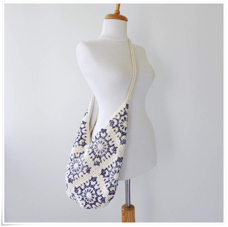 May include: A white crocheted shoulder bag with a blue and white geometric pattern. The bag is hanging on a white mannequin.