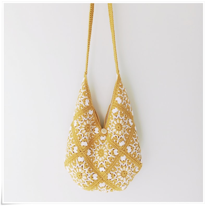 Puede incluir: Un bolso de hombro de crochet amarillo con detalles florales blancos. El bolso tiene una correa larga y un cierre de bot&oacute;n.