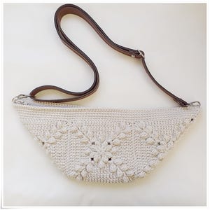 May include: A white crocheted bag with a brown leather strap. The bag has a unique design with a flower pattern.