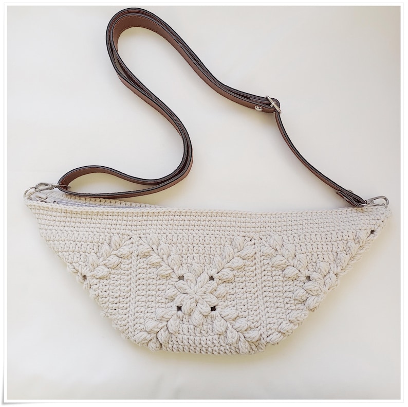 May include: A white crocheted bag with a brown leather strap. The bag has a unique design with a flower pattern.