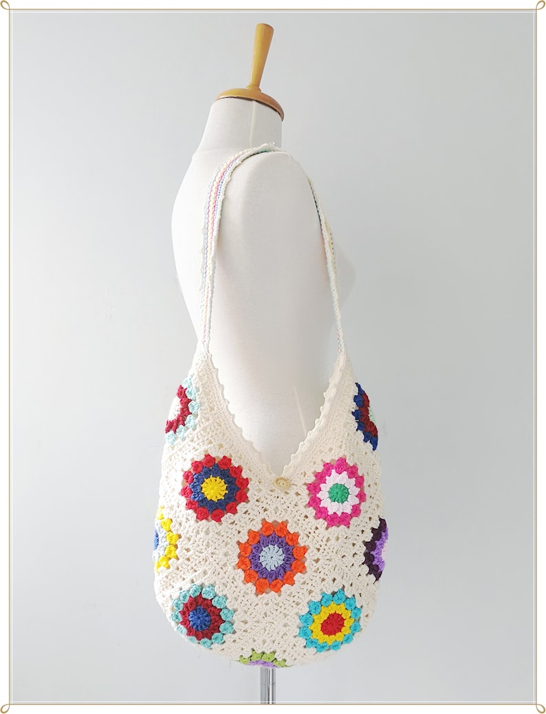 May include: A white crocheted tote bag with a colorful floral design. The bag has a long strap and is made with a granny square pattern.