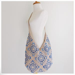 May include: A beige and blue crocheted shoulder bag with a geometric pattern. The bag has a long strap and a wooden button closure.