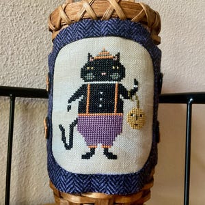 May include: A woven basket with a stitched Halloween design. The design features a black cat wearing a hat and suspenders, holding a pumpkin. The basket has a purple fabric border and wooden handle.