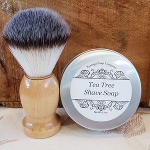 May include: A silver tin of Tea Tree Shave Soap by Tortuga Soap Company. The tin has a decorative design and says "Net Wt. 1.5 oz". A wooden shaving brush with a silver band and black and white bristles is next to the tin.