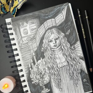 Puede incluir: Un cuaderno de espiral con un dibujo a lápiz en blanco y negro de una mujer sosteniendo un candelabro. El dibujo tiene un tema gótico. Una mariposa decorativa y lápices están junto al cuaderno. También hay una pequeña vela que funciona con pilas.