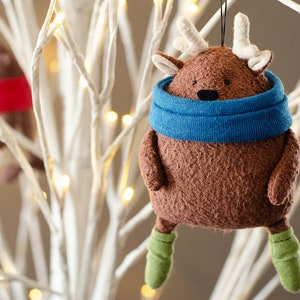 May include: A brown plush reindeer ornament with a blue scarf and green boots hanging on a white branch with white lights.