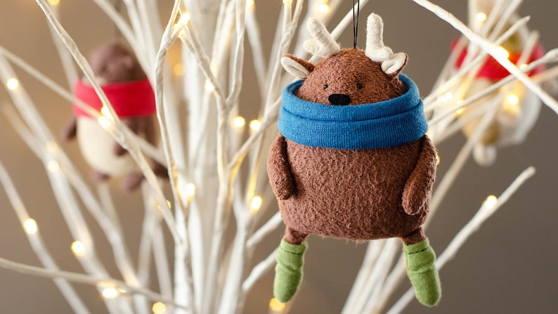 May include: A brown plush reindeer ornament with a blue scarf and green boots hanging on a white branch with white lights.