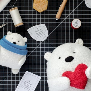 May include: Two white polar bear plush toys, one wearing a blue scarf and the other holding a red heart. The bears are surrounded by sewing supplies, including fabric scraps, thread, scissors, and paper patterns.