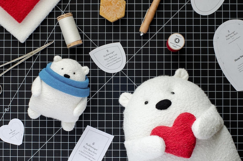 May include: Two white polar bear plush toys, one wearing a blue scarf and the other holding a red heart. The bears are surrounded by sewing supplies, including fabric scraps, thread, scissors, and paper patterns.