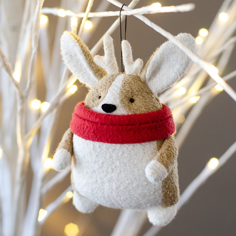 May include: A white and brown plush reindeer ornament with a red scarf. The ornament is hanging from a white branch with white lights.