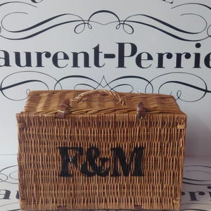May include: A rectangular wicker picnic basket with a handle and leather straps. The basket has the letters "F&M" stenciled in black on the front. The background features the word "CHAMPAGNE" and the name "Laurent-Perrier".