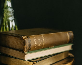 May include: A stack of two antique books with worn, brown leather covers and gold-gilded edges. The top book is titled "Vanity Fair" by W.M. Thackeray. The books rest on a rustic wooden stool, with a blurred vase of flowers in the background.