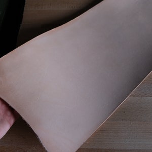 May include: A large piece of light tan leather, held by a hand, with a slightly textured surface. The leather is folded over, revealing its thickness and flexibility. The material appears to be new and ready for crafting.