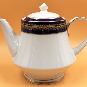 Pode incluir: Bule de porcelana branca com guarnição azul e dourada. A chaleira tem uma tampa com um botão no topo. A chaleira está sobre uma superfície laranja claro.