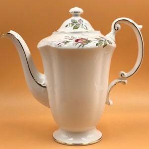 Pode incluir: Uma cafeteira de porcelana branca com acabamento dourado e design floral. A cafeteira tem uma alça curva e uma tampa com um botão.