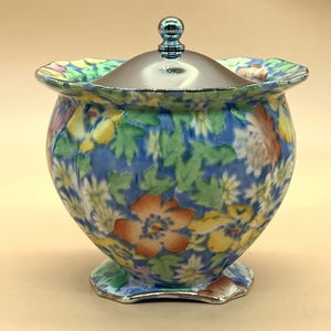 Pode incluir: Um açucareiro de cerâmica com padrão floral azul e branco com tampa prateada. O pote tem uma borda ondulada e uma borda prateada ao redor da base.