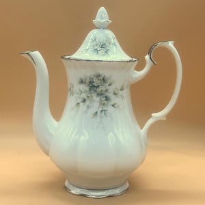 Pode incluir: Cafeteira de porcelana branca com acabamento prateado e design floral. A cafeteira tem um bico curvo, uma alça curva e uma tampa com um botão decorativo. O design floral apresenta flores azuis e verdes.