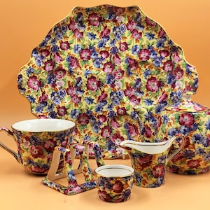 Könnte beinhalten: Ein Vintage-Tee-Set mit einem Blumenmuster. Das Set enthält eine Teekanne, eine Milchkännchen, eine Zuckerdose, eine Tasse und eine Untertasse sowie ein Serviertablett. Das Blumenmuster ist in den Farben Rosa, Lila und Gelb auf weißem Hintergrund.