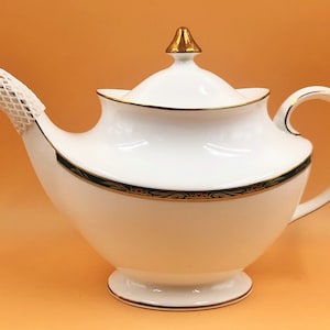 Può includere: Teiera in porcellana bianca con bordo dorato e un motivo floreale verde e oro. La teiera ha un coperchio con un pomo dorato e un manico.