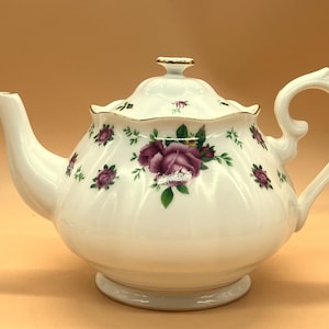 Può includere: Teiera in ceramica bianca con coperchio, beccuccio e manico. La teiera è decorata con rose viola e foglie verdi. I bordi dorati accentuano il coperchio e la base. La teiera è progettata per servire il tè.