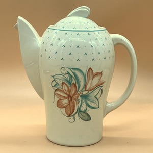 Puede incluir: Cafetera de cerámica blanca con un diseño alto y esbelto. Presenta un motivo floral en tonos marrón, verde azulado y verde. La tapa y el borde tienen un patrón de puntos verde azulado. El pico es largo y angular, y el asa es curva.