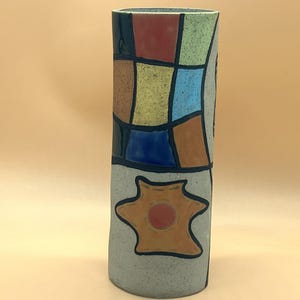 Puede incluir: Un jarrón cilíndrico de cerámica con un diseño de patchwork. La parte superior presenta un mosaico de cuadrados rojos, verdes, amarillos, azules y marrones. La sección inferior tiene una forma de estrella con un centro naranja. El jarrón mide unos 30 cm de alto.
