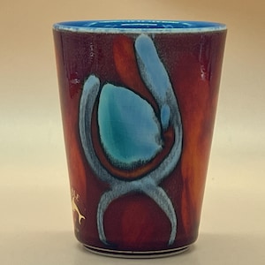 Könnte beinhalten: Ein Keramikbecher mit lebendigem Design. Die Außenseite hat eine tiefrote Basis mit orangefarbenen und blauen Akzenten. Eine große, abstrakte blaue Form ist auf die Vorderseite gemalt. Der Rand ist hellblau.