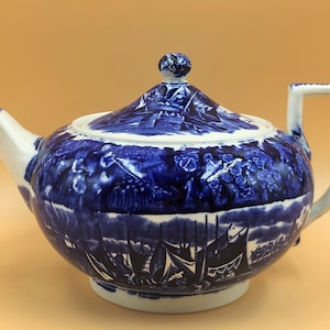 May include: A blue and white ceramic teapot with a lid and handle. The teapot features a detailed landscape design with ships and buildings. The spout and handle are white. The teapot is a classic design.