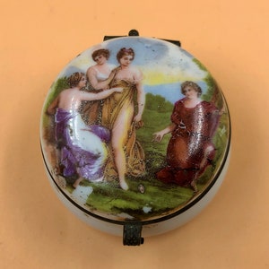 Könnte beinhalten: Eine runde Porzellan-Schmuckdose mit einem Scharnierdeckel. Der Deckel zeigt eine handgemalte Szene mit drei Frauen in fließenden Roben vor einem Landschaftshintergrund. Die Dose ist mit einem goldenen Rand verziert.
