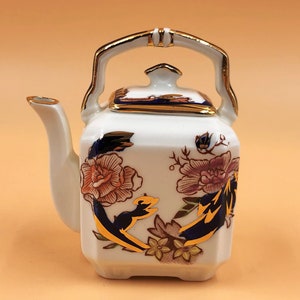 May include: A white porcelain teapot with a square body, featuring floral designs in shades of orange, burgundy, and gold. The teapot has a gold-trimmed handle, spout, and lid, and is approximately 5 inches tall.