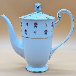 Pode incluir: Uma chaleira de porcelana branca com uma borda dourada e um padrão floral de rosas cor-de-rosa. A chaleira tem uma tampa com um botão no topo e uma alça no lado.