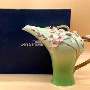May include: A decorative ceramic pitcher with a light green and brown handle. The pitcher is adorned with pink cherry blossom accents and the text "Dan Samuels". The pitcher is next to a dark blue box.