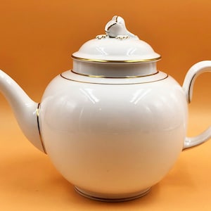 Pode incluir: Bule de chá de porcelana branca com acabamento dourado e tampa em forma de sapo. A chaleira tem um corpo redondo e um bico curvo.