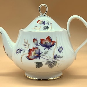 以下が含まれることがあります： 赤と紫の花、緑の葉、金の装飾が施された、白い磁器のティーポット。 ティーポットは、湾曲したハンドルと注ぎ口、蓋にループが付いています。