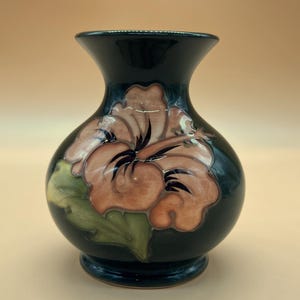 May include: A dark green ceramic vase with a glossy finish, featuring a hand-painted floral design in shades of soft peach, light brown, and muted green. The vase has a rounded body, a flared neck, and a circular base, showcasing a smooth surface.