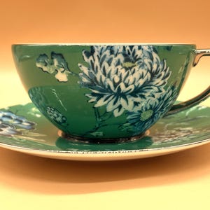 May include: A teal teacup and saucer set featuring a white and dark blue chrysanthemum floral pattern. The teacup has a metallic silver handle and a matching silver rim on both the cup and the saucer, providing a reflective finish to the set.