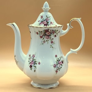 May include: White porcelain coffee pot with a floral rose pattern in pink and purple. The pot has a curved spout, a curved handle, and a lid with a decorative finial. Gold trim accents the handle and base.