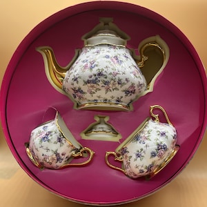 Puede incluir: Un juego de té de porcelana blanca con un patrón floral de flores rosas, moradas y azules, con bordes dorados en la tetera, la tapa, la lechera y el azucarero. El conjunto se presenta en una caja circular rosa con compartimentos moldeados.