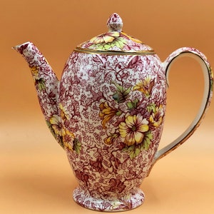 Pode incluir: Uma chaleira de cerâmica branca com um padrão floral em tons de vermelho, amarelo e verde. A chaleira tem uma borda dourada em torno da tampa e do bico.