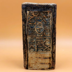 Puede incluir: Un jarrón rectangular de cerámica con un esmalte marrón y azul. El jarrón tiene un patrón geométrico tallado en la superficie.