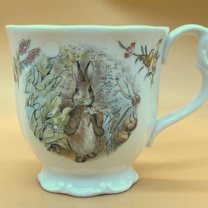 Puede incluir: Taza de té de cerámica blanca con una ilustración detallada de un conejo en un jardín. La taza presenta un asa decorativa y una base festoneada. La obra de arte incluye varias plantas y flores, con una paleta de colores suaves.