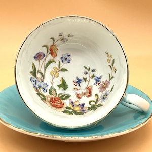 Puede incluir: Juego de taza y platillo blancos con diseño floral. La taza tiene un borde dorado y un asa. El platillo es de color azul turquesa claro con borde dorado. La taza y el platillo están decorados con flores y mariposas coloridas.