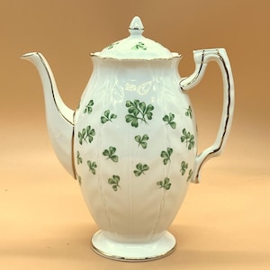 May include: White ceramic teapot with a lid, handle, and spout. The teapot is decorated with green shamrock designs and gold trim. The teapot is approximately 10 inches tall.