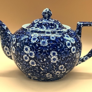 May include: A ceramic teapot featuring a deep blue background with a dense, repeating white floral pattern. The teapot includes a matching lid with a small knob, a curved spout, and a sturdy handle, all covered in the same intricate botanical design.
