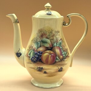Puede incluir: Una tetera de porcelana de color crema con un motivo de frutas pintado a mano en la parte frontal. El diseño incluye un melocotón, uvas y hojas, con bordes dorados en el asa, el pico y la tapa. La tapa cuenta con un pequeño remate superior.