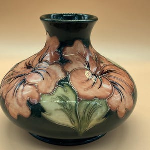 May include: A bulbous ceramic vase featuring a dark green glossy glaze with raised relief floral motifs in shades of peach and light green. The vase has a wide, rounded body that tapers into a narrow, flared neck with a smooth, reflective finish.