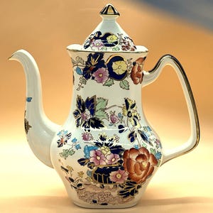 Pode incluir: Cafeteira de cerâmica branca com tampa, apresentando um design floral em tons de azul, rosa e laranja. A alça e o bico são brancos com detalhes dourados. A cafeteira tem uma forma alta e esguia e uma tampa decorativa com um botão dourado.