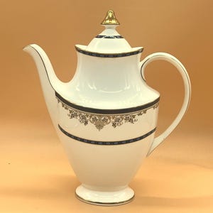 Pode incluir: Cafeteira de porcelana branca com um botão e alça dourados. A cafeteira apresenta uma faixa decorativa de detalhes dourados e azul marinho ao redor do corpo e da tampa. Um design clássico para servir café ou chá.